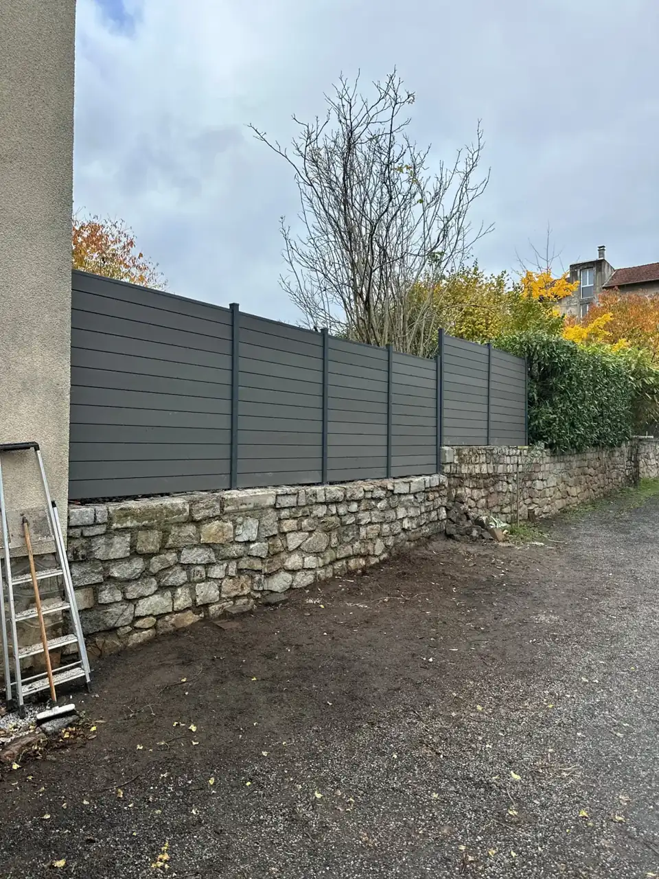 Clôture en panneaux de bois gris avec mur en pierre sous un ciel nuageux.
